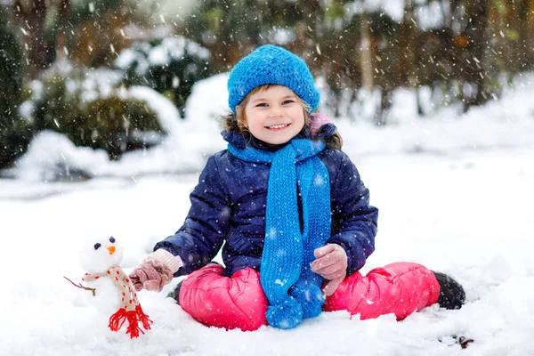 Söt liten småbarn flicka gör mini snögubbe och äter morot näsa. Underbar frisk glad barn leka och ha kul med snö, utomhus på kall dag. Aktiv fritid med barn på vintern — Stockfoto