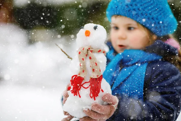 かわいい小さな幼児の女の子はミニ雪だるまを作り、ニンジンの鼻を食べる。寒い日には屋外で雪と遊ぶと楽しい健康的な幸せな子供。冬の子供たちとのアクティブなレジャー — ストック写真