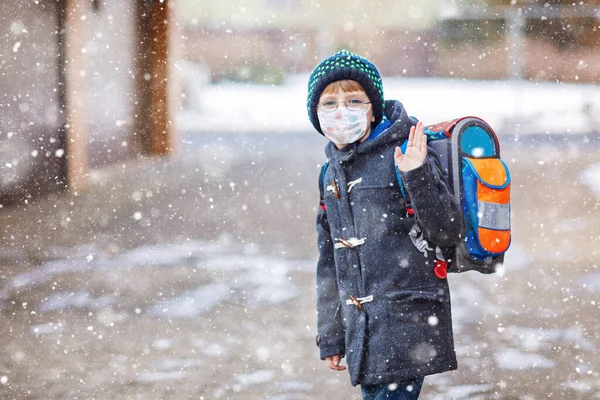 一个戴眼镜、戴着医疗面罩的小男孩在关了门后去学校的路上。儿童背包包。冬日的小学生穿着保暖的衣服.日冕大流行病期间的检疫时间 — 图库照片