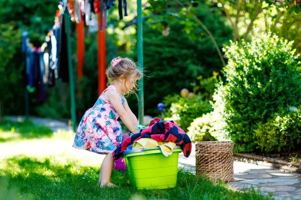 Gadis penghibur menggantung pakaian, gaun bersih segar dan celana untuk pengeringan di taman, luar ruangan setelah membuat cucian. Anak bahagia membantu dalam rumah tangga. Keluarga bekerja sama, anak belajar untuk membantu. — Stok Foto