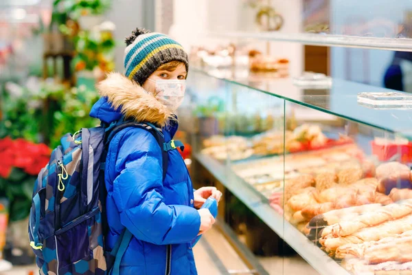 Een jongen met een medisch masker koopt brood en gebak voor de schoollunch in de bakkerij. Kind met rugzak en winterkleding. Schoolkind tijdens vergrendeling en quarantaine tijdens corona pandemische ziekte — Stockfoto