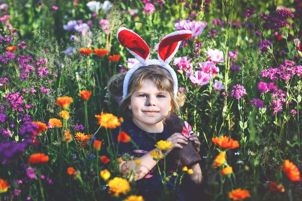 可爱可爱迷人的小女孩，长着复活节兔子的耳朵，在花草甸里吃巧克力兔子的肖像。在阳光灿烂的日子，带着五彩缤纷的花朵，在户外微笑着快乐的孩子. — 图库照片