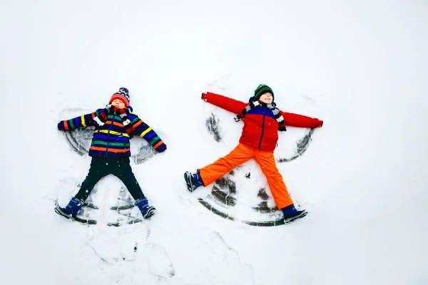 Due fratellini bambini ragazzi in abiti invernali colorati facendo angelo della neve, sdraiati sulla neve. Tempo libero attivo all'aperto con i bambini in inverno. Fratelli felici con cappello caldo, guanti, moda invernale. — Foto Stock