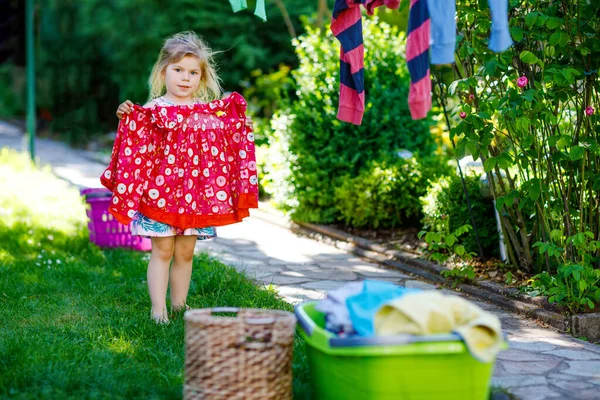 Kleinkind-Mädchen hängende Kleidung, frische saubere Kleider und Hosen zum Trocknen im Garten, im Freien nach dem Waschen. Glückliches Kind, das im Haushalt hilft. Familie arbeitet zusammen, Kind lernt zu helfen. — Stockfoto