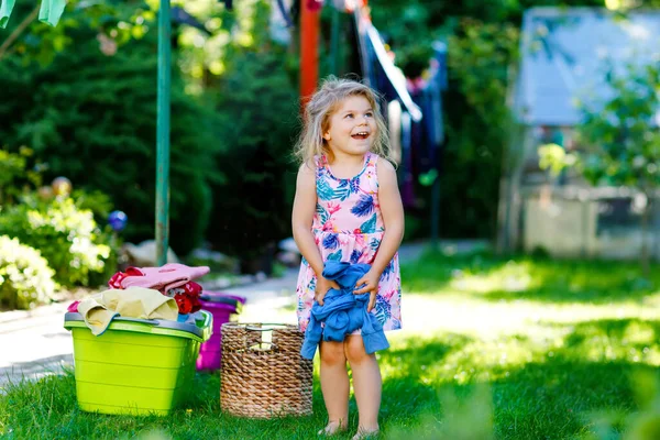 Batole dívka visí oblečení, čerstvé čisté šaty a kalhoty pro sušení v zahradě, venku po výrobě prádla. Šťastné dítě pomáhá v domácnosti. Rodina spolupracuje, dítě se učí pomáhat. — Stock fotografie