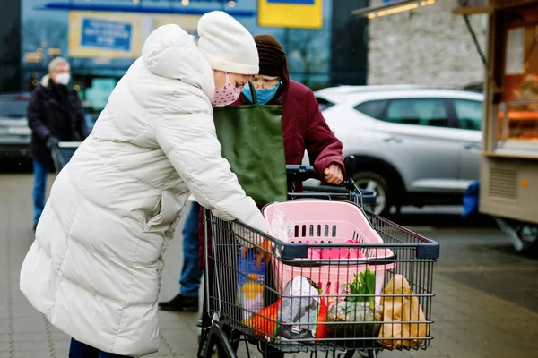 Seniorin und Sozialarbeiterin mit medizinischer Maske wegen Coronavirus-Pandemie. Tochter oder Enkelin helfen Großmutter beim Einkaufen im Supermarkt, schieben Einkaufswagen mit Lebensmitteln, im Freien — Stockfoto