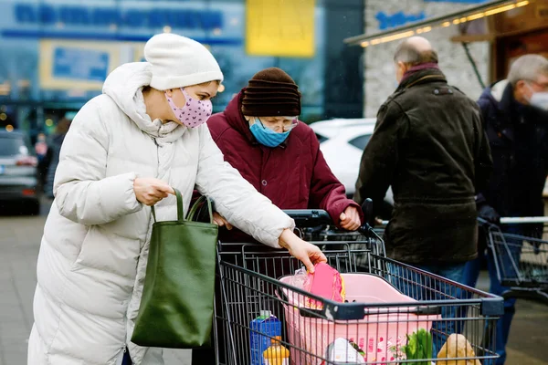 Senior woman and social worker with medical mask due pandemic coronavirus disease. Daughter or granddaughter help grandmother with shopping in supermarket, push cart trolley with foods, outdoors — Stock Photo, Image