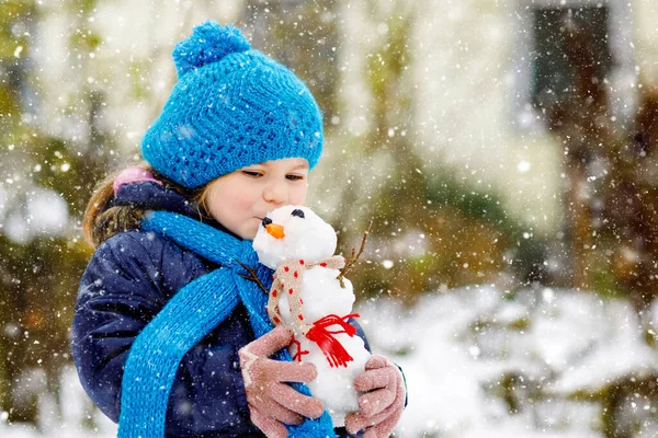 Menina pequena bonito fazendo mini boneco de neve e comer nariz de cenoura. Adorável criança feliz saudável brincando e se divertindo com neve, ao ar livre no dia frio. Lazer ativo com crianças no inverno — Fotografia de Stock