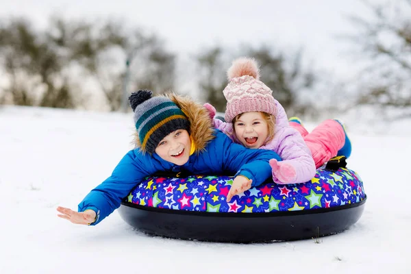 Aktív kisbaba és iskolás fiú együtt csúsznak le a hegyről hócsővel. Boldog gyerekek, testvérek, akik télen a szabadban szórakoznak szánkón. Testvérek csövek havas lefelé, családi idő. — Stock Fotó