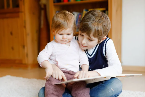 Menino da escola lendo livro para a menina da criança, dois irmãos sentados juntos e ler livros. Linda família adorável no amor, bebê bonito e criança se divertindo em casa, dentro de casa. — Fotografia de Stock