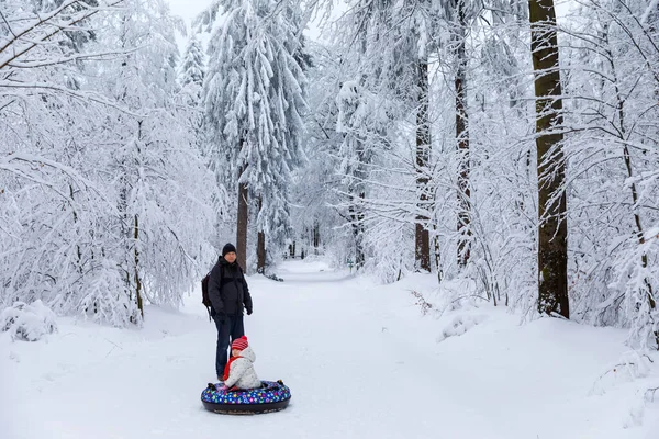 年轻的父亲把小女孩拖在雪地管子上。可爱的小快乐的孩子在冬天穿着五彩缤纷的轮胎在户外玩.家庭、女儿和男人在外面雪地的森林里徒步旅行和散步. — 图库照片