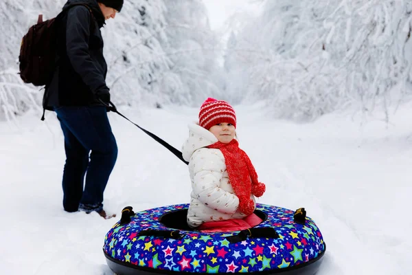 Genç baba, küçük kızı kar tüpüne bindirdi. Sevimli küçük mutlu çocuk renkli lastiklerle kışın dışarıda eğleniyor. Aile, kız ve adam karlı ormanda yürüyüş yapıyor ve yürüyor.. — Stok fotoğraf
