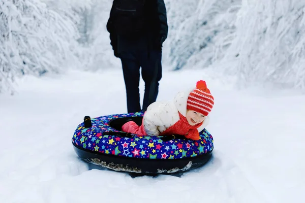 El padre joven tira de la niña en el tubo de nieve. Lindo niño feliz que se divierte al aire libre en invierno en el neumático colorido. Familia, hija y hombre senderismo y caminar en el bosque nevado, al aire libre. — Foto de Stock