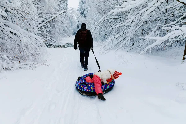 年轻的父亲把小女孩拖在雪地管子上。可爱的小快乐的孩子在冬天穿着五彩缤纷的轮胎在户外玩.家庭、女儿和男人在外面雪地的森林里徒步旅行和散步. — 图库照片
