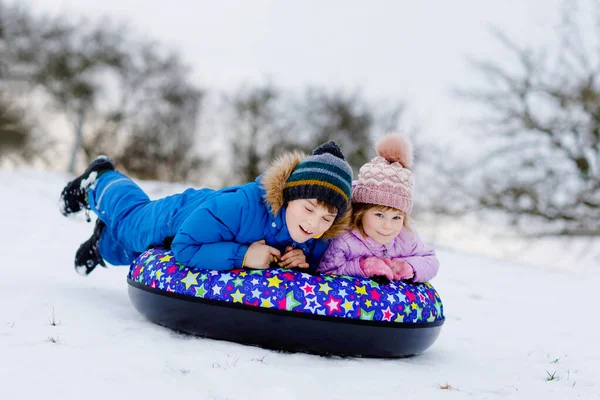 Actieve peuter meisje en schooljongen glijden samen de heuvel af op sneeuw buis. Vrolijke kinderen, broers en zussen die in de winter plezier hebben op slee. Broer en zus tubing besneeuwde afdaling, familie tijd. — Stockfoto