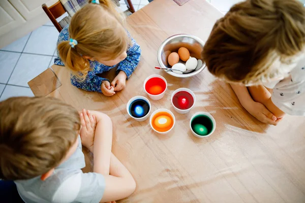 Excited little toddler girl and two kids boys coloring eggs for Easter. Three children, siblings looking surprised at colorful eggs, celebrating holiday with family. From above, unrecognized faces — Stock Photo, Image