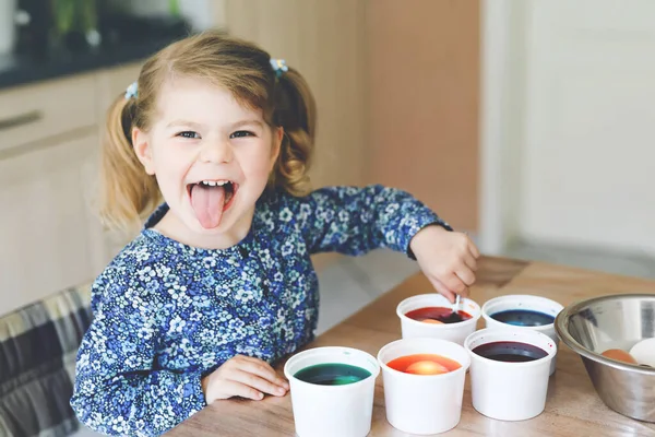 Excited little toddler girl coloring eggs for Easter. Cute happy child looking surprised at colorful colored eggs, celebrating holiday with family. Adorable kid at home with different bright colors. — Stock Photo, Image