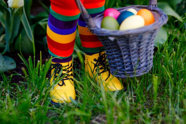 Close-up van de benen van peuter meisje met kleurrijke kousen en schoenen en mand met gekleurde eieren. Kind heeft plezier met traditionele paaseieren jacht, buiten. Viering van christelijke feestdagen. — Stockfoto