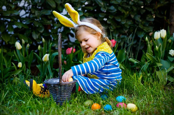 Söt liten småbarn flicka med kaninöron har kul med traditionella påskägg jakt på varm solig dag, utomhus. Glada barn firar semester. Baby hitta, färgade ägg i trädgården — Stockfoto