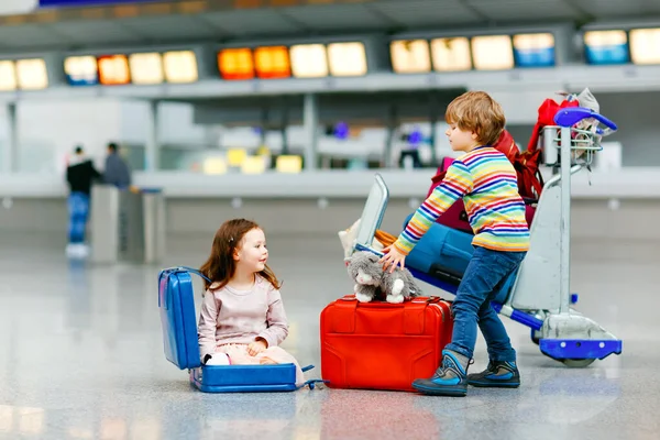 Ragazzina e ragazzo carino all'aeroporto. Bambini stanchi, fratelli seduti a terra in attesa di un volo ritardato. Bambini che giocano insieme con il carrello bagagli con valigie al terminal. Stile di vita di viaggio — Foto Stock