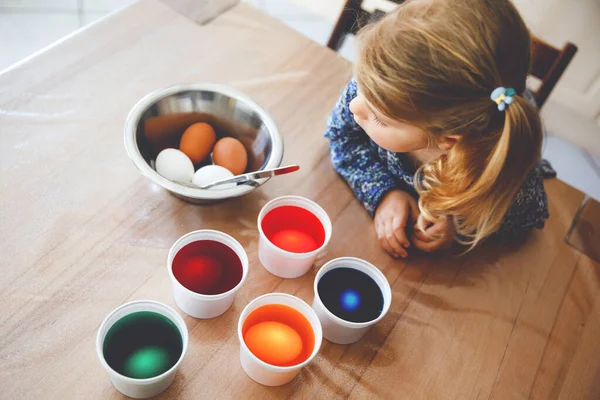 イースターのために卵を着色する興奮した小さな幼児の女の子。カラフルな色の卵を見てかわいい幸せな子供は、家族と一緒に休日を祝います。上から顔が見えない. — ストック写真