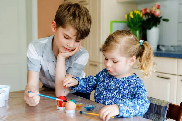 Nadšený malý batole holka a starší bratr, školák zbarvení vejce na Velikonoce. Dvě roztomilé děti, sourozenci vypadající překvapeni barevnými vejci, slaví dovolenou s rodinou. — Stock fotografie