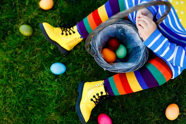 Närbild av ben av småbarn flicka med färgglada strumpor och skor och korg med färgade ägg. Barn har roligt med traditionella påskägg jakt, utomhus. Oigenkännligt ansikte, inget ansikte. — Stockfoto