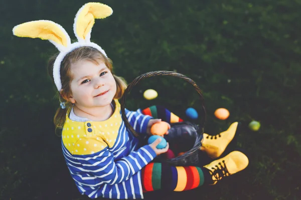 Roztomilé malé batole dívka sedí na zelené trávě a nosí králičí uši, baví se s tradiční velikonoční barevné vejce lovit na teplý slunečný den, venku. Šťastné dítě slaví rodinnou dovolenou. — Stock fotografie