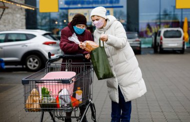 Kıdemli kadın ve tıbbi maskeli sosyal hizmetler görevlisi pandemik koronavirüs hastalığı nedeniyle. Kızı ya da torunu süpermarkette alışveriş yaparken büyükannesine yardım ediyor, market arabasında yiyecek satıyor, açık havada geziyor.