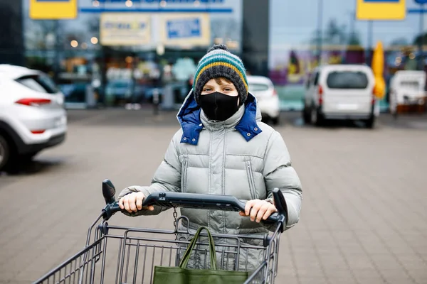Kid boy wear medical mask as protection against pandemic coronavirus disease. Child use protective equipment against covid 19 and push cart trolley for shopping in supermarket, outdoors, winter time. — Stock Photo, Image