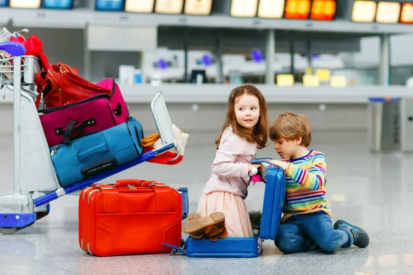 Aranyos kislány és fiú a reptéren. Fáradt gyerekek, testvérek ülnek a földön, és a késleltetett repülésre várnak. A gyerekek együtt játszanak a bőröndökkel a terminálnál. Utazási életmód — Stock Fotó