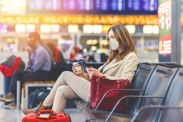 Een vrouw met een medisch masker die boek leest en koffie drinkt. Vrouw overstuur door vlucht annulering, zittend in bijna lege internationale luchthaven terminal als gevolg van coronavirus pandemie reisbeperkingen Stockfoto