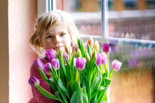 Little girl sitting by window with tulip flowers bouquet. Happy child, indoors. Mothers day, valentines day or birthday concept. — Stock Photo, Image