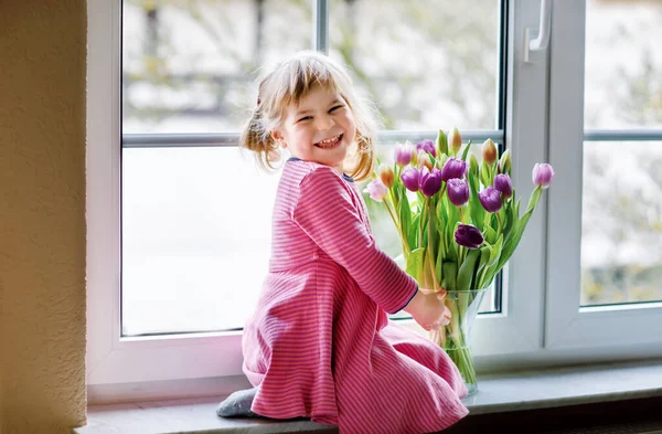 チューリップの花の花束を窓のそばに座っている少女。幸せな子供室内で。母の日、バレンタインデー、誕生日のコンセプト. — ストック写真