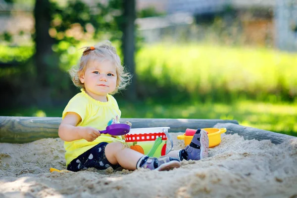 Schattig peuter meisje spelen in zand op buiten speeltuin. Mooie baby die plezier heeft op zonnige warme zomerse zonnige dag. Gelukkig gezond kind met zand speelgoed en in kleurrijke mode kleding. — Stockfoto