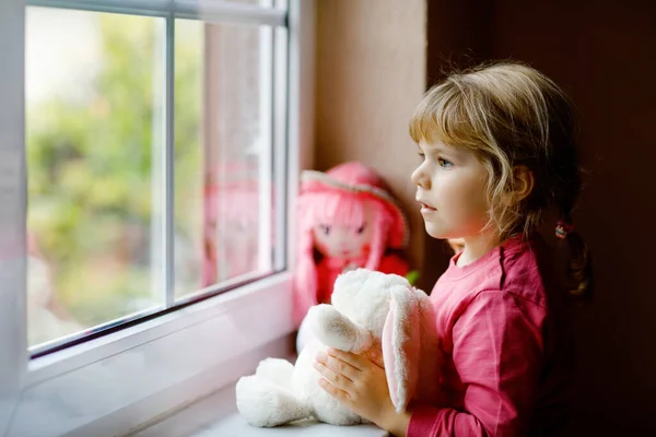 かわいい幼児の女の子は窓のそばに座って雨の日を見ています。人形と柔らかいおもちゃの幸せな気分で子供を夢見ています。コロナウイルスパンデミック時の自己分離概念。孤独な子. — ストック写真
