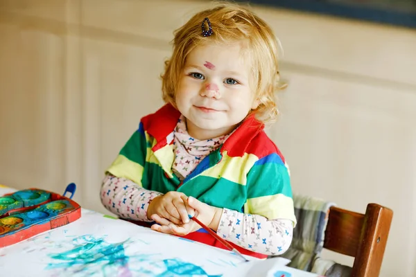 Linda niña adorable aprendiendo pintura con acuarelas. Pequeño niño dibujando en casa, usando pinceles de colores. Saludable hija feliz experimentando con colores, agua en casa o vivero. — Foto de Stock