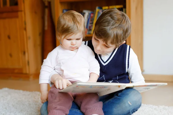 En skolegutt som leser bok for småbarnsjente, to søsken som sitter sammen og leser bøker. Vakker familie i kjærlighet, søt baby og barn som har det gøy hjemme, inne. – stockfoto