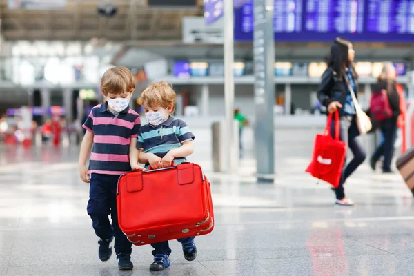 Due fratellini indossano una maschera medica e vanno in vacanza con la valigia all'aeroporto, al chiuso. Fratelli gemelli felici che camminano per il check-in o l'imbarco per il volo durante la pandemia del virus della corona — Foto Stock