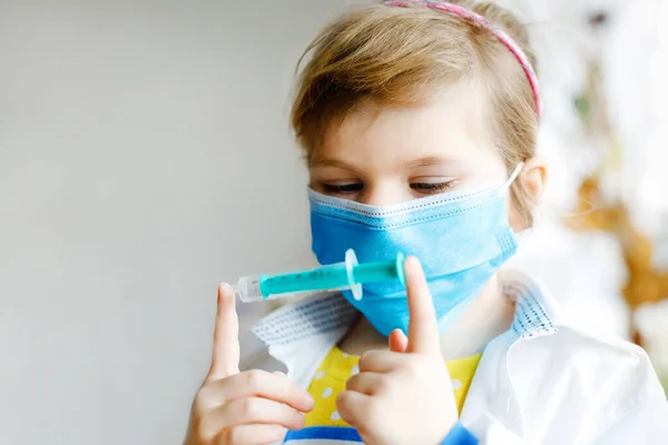 Маленькая девочка делает укол. Симпатичный ребенок в медицинской маске, играет в доктора, держит шприц с вакциной. Концепция коронавирусной ковидовой вакцинации. Детские ролевые игры дома. — стоковое фото
