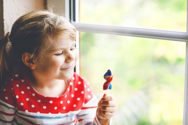 Cute little toddler girl sitting by window on rainy day during pandemic coronavirus quarantine. Adorable child eating and licking rainbow colored lollipop — Stock Photo, Image