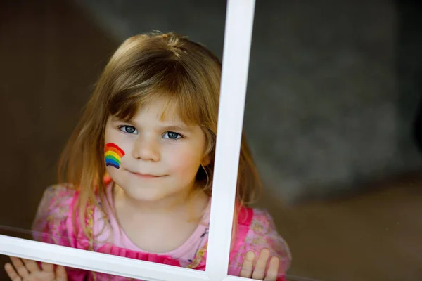 Carino solitario bambina in abito da principessa seduta alla finestra con arcobaleno con colori colorati sul viso durante la quarantena pandemica coronavirus. I bambini fanno e dipingono arcobaleni in tutto il mondo — Foto Stock