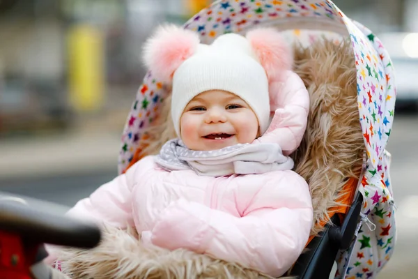 Linda niña hermosa sentada en el cochecito o cochecito en el frío otoño, invierno o día de primavera. Feliz niño sonriente en ropa de abrigo, abrigo de bebé con estilo de moda y sombrero. Nieve cayendo — Foto de Stock