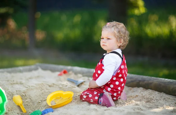 Menina bonito brincando na areia no parque infantil ao ar livre. Bebê bonito em calças de goma vermelha se divertindo no dia de verão quente ensolarado. Criança com brinquedos coloridos de areia. Bebê ativo saudável ao ar livre joga jogos — Fotografia de Stock
