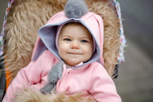 Schattig klein mooi babymeisje zittend in de kinderwagen of kinderwagen op herfstdag. Gelukkig lachend kind in warme kleren, mode stijlvolle roze babyjas met konijnenoren. Baby gaat wandelen met ouders. — Stockfoto