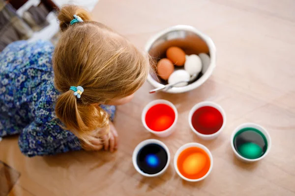 Excited little toddler girl coloring eggs for Easter. Cute happy child looking surprised at colorful colored eggs, celebrating holiday with family. From above, unrecognized face. — Stock Photo, Image