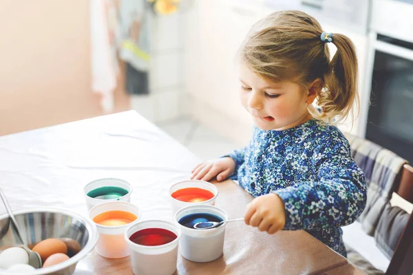Excited little toddler girl coloring eggs for Easter. Cute happy child looking surprised at colorful colored eggs, celebrating holiday with family. Adorable kid at home with different bright colors. — Stock Photo, Image