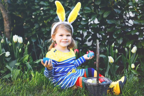 Söt liten småbarn flicka med kaninöron har kul med traditionella påskägg jakt på varm solig dag, utomhus. Glad barn firar familj kristna semester med korg med färgade ägg — Stockfoto