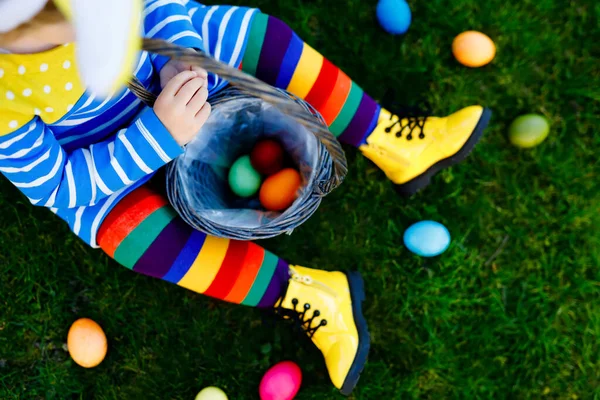 Close-up of legs of toddler girl with colorful stockings and shoes and basket with colored eggs. Child having fun with traditional Easter eggs hunt, outdoors. Unrecognizable face, no face. — Stock Photo, Image
