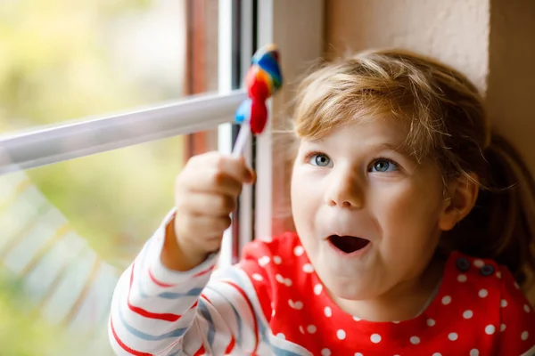 Carina bambina seduta vicino alla finestra nei giorni di pioggia durante la quarantena del coronavirus pandemico. Adorabile bambino mangiare e leccare lecca lecca lecca-lecca color arcobaleno — Foto Stock